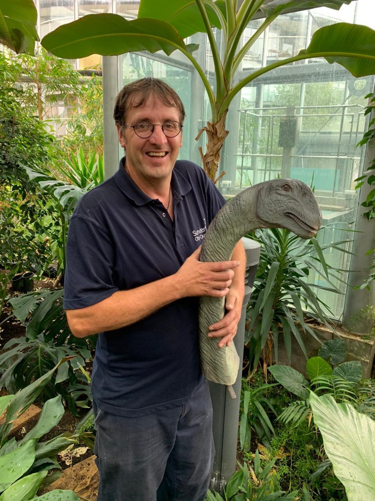 Man standing in an indoor tropical environment, carrying a model dinosaur head